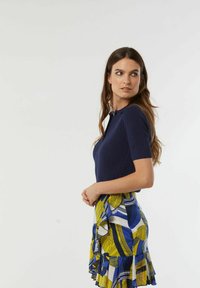 Navy gebreid top met korte mouwen, gecombineerd met een multicolore patroonrok met gele, blauwe en witte kleuren, versierd met ruffles.