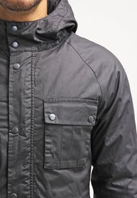 Ciemnoszara kurtka z kapturem, wyposażona w zapięcie na zatrzask, teksturowaną tkaninę oraz kieszeń na klapę z ozdobnym przeszyciem i zatrzaskami.