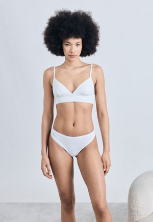 Femme aux cheveux afro naturels se tenant face à l'avant, portant un soutien-gorge blanc et un sous-vêtement assorti blanc, sur un fond uni.