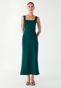 Tyrkysové maxi šaty se čtvercovým výstřihem, přiléhavým tělem a splývavou sukní. Hladká textura látky. Modelka má na sobě černé ploché sandály.