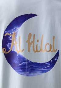 Gaiši zilā krāsā t-krekls ar lielu purpura mēness dizainu un mākoņu rakstiem, kā arī tekstu "Al Hilal" metāliskā zelta burtiem.
