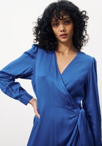 Blaues Wickelkleid mit langen Ärmeln, weicher Textur, tiefem V-Ausschnitt und Seitenschleifen-Detail. Verfügt über geraffte Schultern für ein zusätzliches Design.