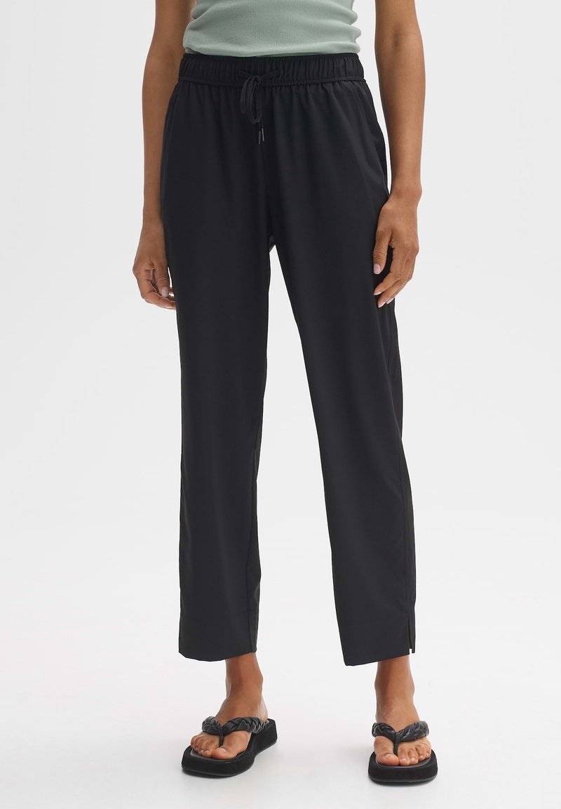 Femme portant un pantalon noir à cordon de serrage et des sandales à plateforme noires tressées, debout devant un fond clair uni.