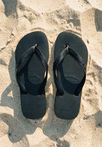 Zwarte Havaianas slippers rusten op gestructureerd zand en werpen duidelijke schaduwen in fel zonlicht.