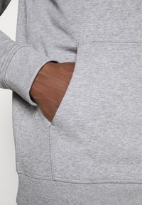 Main à la peau foncée glissée dans la poche avant d'un sweat-shirt gris clair avec poignets et ourlet côtelés.