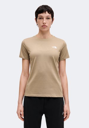 Mujer con cabello corto y oscuro vestida con una camiseta beige de The North Face y pantalones negros, de pie frente a un fondo gris claro.