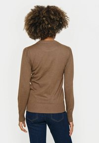 Brauner Langarmpullover mit geripptem Halsausschnitt und Bündchen, glatter Textur, figurbetontem Schnitt und subtil gepufften Schultern. Getragen mit dunklen Jeans.
