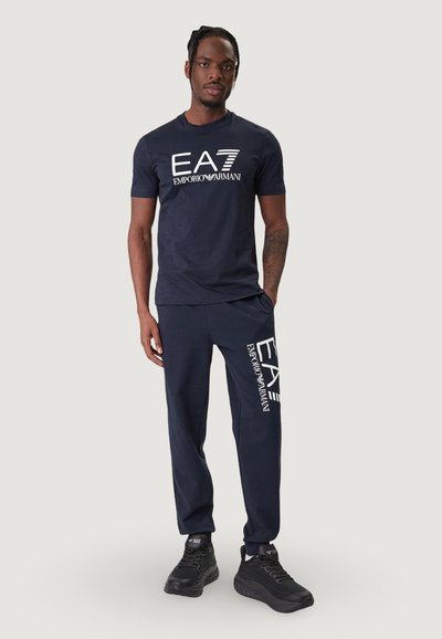 Тъмносин памучен T-shirt и съвпадащи панталони, с бяло лого "EA7 Emporio Armani" отпред и отстрани, с просторна кройка и гладка текстура.