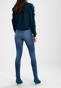 Türkiser Pullover mit Rüschendetails an den Ärmeln, kombiniert mit blauen Skinny-Jeans. Das Modell trägt türkise Ankle Boots. Einfaches, lässiges Design.