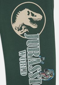 Tessuto di cotone verde con una silhouette di T-rex beige e "JURASSIC WORLD" in bianco, arricchito da un dettaglio grafico di un dinosauro blu.