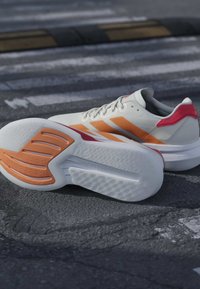 Chaussures de sport blanches et orange avec des semelles texturées reposant sur une route asphaltée avec des marquages de passage piéton.