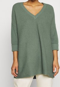 Maglione verde lavorato a maglia con scollo a V, maniche a tre quarti e vestibilità ampia. Il tessuto strutturato presenta un motivo a nido d'ape.