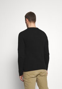 Schwarzer Pullover mit langen Ärmeln, strukturiertem Strickmuster und rundem Ausschnitt, kombiniert mit beigefarbenen Hosen. Die Rückansicht zeigt ein tailliertes Design.