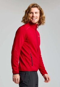 Homme aux cheveux bouclés souriant, portant une veste zippée rouge et un pantalon noir, se tenant devant un fond gris uni.