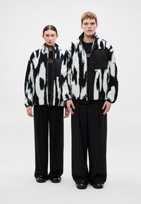 Fekete-fehér ikat mintás polár dzsekik kontrasztos fekete zsebekkel, széles fekete nadrággal és fényes lábbelivel párosítva.