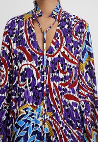 Blouse multicolore présentant des motifs violets, rouges et bleus, un col montant et une patte de boutonnage à l'avant. Confectionnée dans un tissu léger à texture douce.