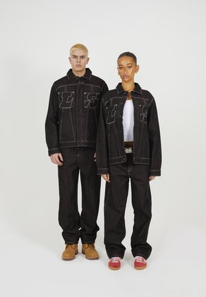 Jonge man en vrouw staand, gekleed in zwarte denimjacks en broeken met grote gestikte letters, witte blouse en casual schoenen.