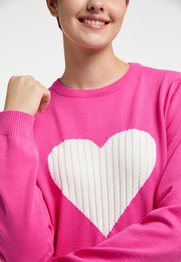 Pulover tricotat roz, cu un design mare de inimă albă cu nervuri pe față. Puloverul are un guler rotund și manșete cu nervuri.