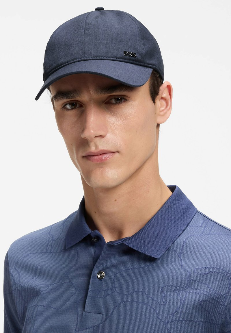 Jeune homme portant une casquette bleu marine avec le logo "BOSS" et un polo bleu assorti à col avec un motif subtil, regardant vers l'avant.