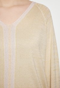Pulover texturat de un aurie deschis, cu decolteu �în V, strălucire subtilă și un design cu nervuri orizontale și accente modelate de-a lungul gulerului.