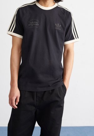 Mężczyzna w czarnej koszulce Adidas Originals z beżowymi obszyciami i trzema paskami na rękawach, z rękami w kieszeniach, na jednolitym tle.