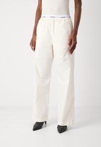 White, lightweight, elastic waist pants with a smooth texture, wide-leg design, and a branded waistband. Worn with black pointed heels.