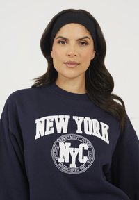 Donna con capelli scuri e mossi che indossa una fascia nera e una felpa blu navy con il testo bianco "NEW YORK NYC" su uno sfondo chiaro e semplice.
