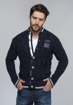 Marinefarbener Cardigan mit Kabelstrick-Design, weißen Akzenten am Kragen und an den Ärmeln, großen Knöpfen und einem gestickten Logo auf der Brust.