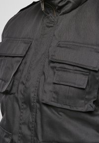 Černá užitková bunda z odolné látky, vybavená několika předními kapsami a zapínáním na zip. Texturovaný povrch se strukturovaným střihem.
