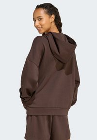Donna che sorride girando la testa, indossando una felpa oversize marrone scuro e pantaloni corti abbinati su uno sfondo semplice.