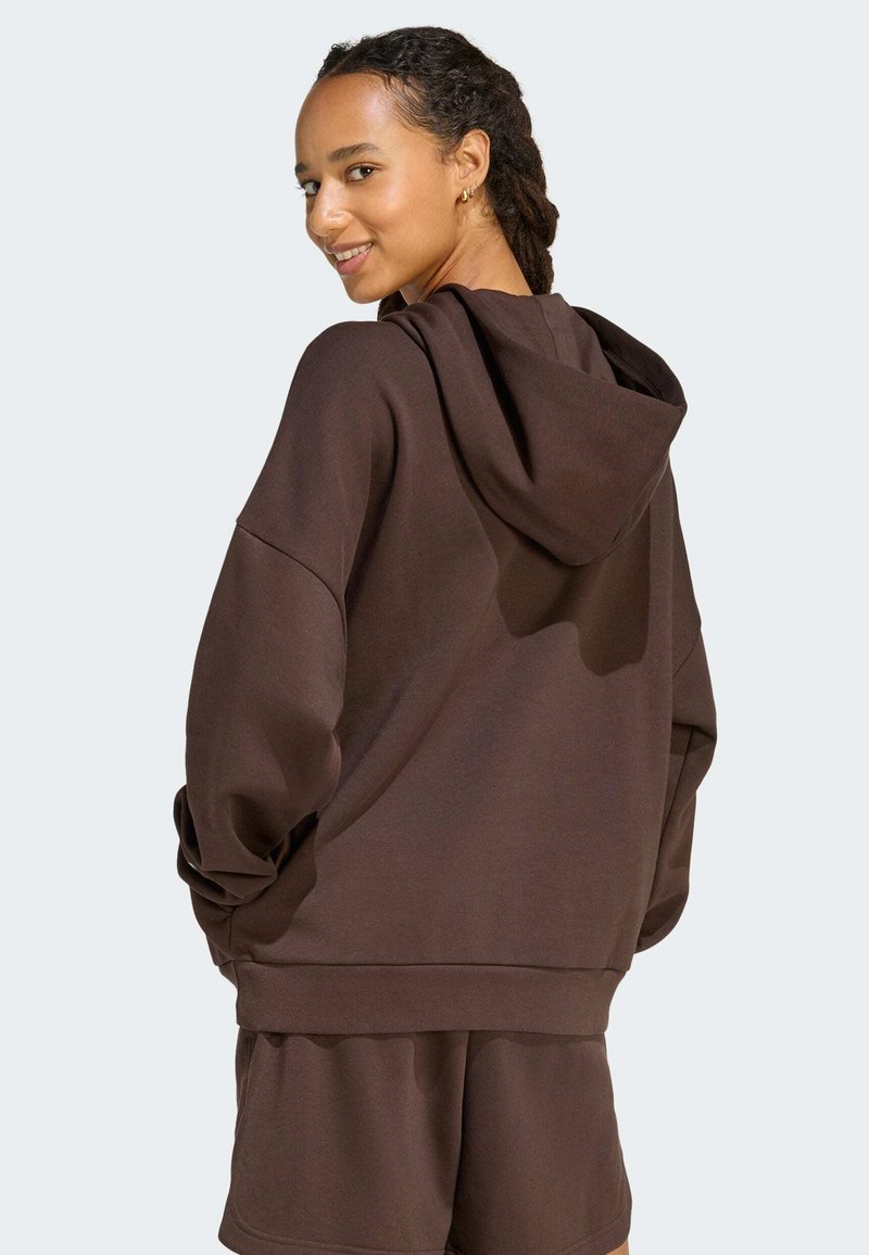 Femme souriante par-dessus l'épaule, portant un sweat à capuche oversized marron foncé et un short assorti, sur fond uni.