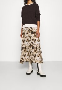 Dunkelbrauner, strukturierter Strickpullover über einem cremefarbenen Oberteil; fließender, gemusterter Rock in Beige mit braunen Akzenten; weiße Stiefeletten.