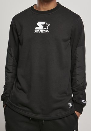 Homme portant un sweat-shirt noir à manches longues Starter avec un logo blanc sur la poitrine et un patch sur le bas de la manche, mains dans les poches d'un pantalon noir.