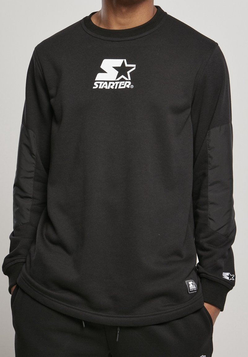 Homme portant un sweat-shirt noir à manches longues Starter avec un logo blanc sur la poitrine et un patch sur le bas de la manche, mains dans les poches d'un pantalon noir.