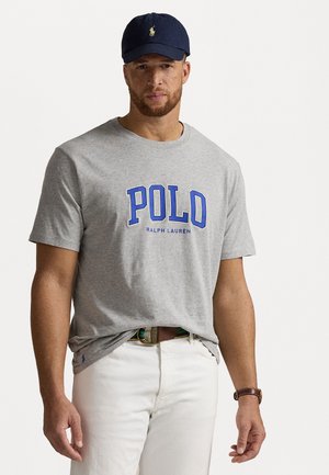 Grå bomulds t-shirt med en blå "POLO" grafik og "RALPH LAUREN" tekst. Kombineret med hvide bukser og en marineblå baseballkasket.