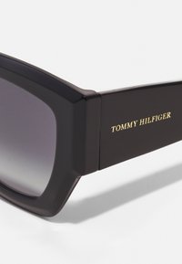 Ochelari de soare negri, cu formă rectangulară, dotati cu lentile gri în degrade, un cadru gros și branding auriu "TOMMY HILFIGER" pe lateral.
