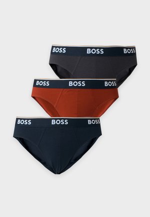 Noormees seisab paljajalu, kandes roostepunaseid bokse, millel on must vöörihm ja sildi "BOSS", ühetaolise heledalt halli tausta ees.