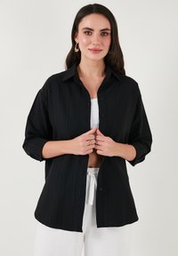 Black button-up shirt with a textured fabric. It has a collar, long sleeves rolled up, and a relaxed fit, paired with white pants.