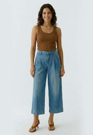 Femme debout portant un haut marron sans manches, un jean bleu à jambes larges et courtes, et des talons marron ouverts devant un fond clair uni.