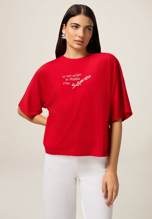 T-shirt rouge à manches courtes en coton, avec un texte en italique blanc brodé, associé à un pantalon blanc. Texture lisse, coupe décontractée.