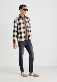 Camisa de franela a cuadros en crema, azul marino y marrón, llevada sobre una camiseta marrón. Jeans oscuros y zapatillas gris claro con detalles en blanco completan el look.