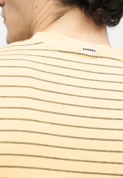 Άνδρας που φοράει ανοιχτό κίτρινο T-shirt με λεπτές οριζόντιες μαύρες ρίγες και μικρή ετικέτα "SANDRO" στο πάνω μέρος της πλάτης κοντά στο γιακά.