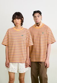 Två individer bär löst sittande, kortärmade randiga t-shirts i gula, orange och lila nyanser med en grön logotyp på vänster sida.