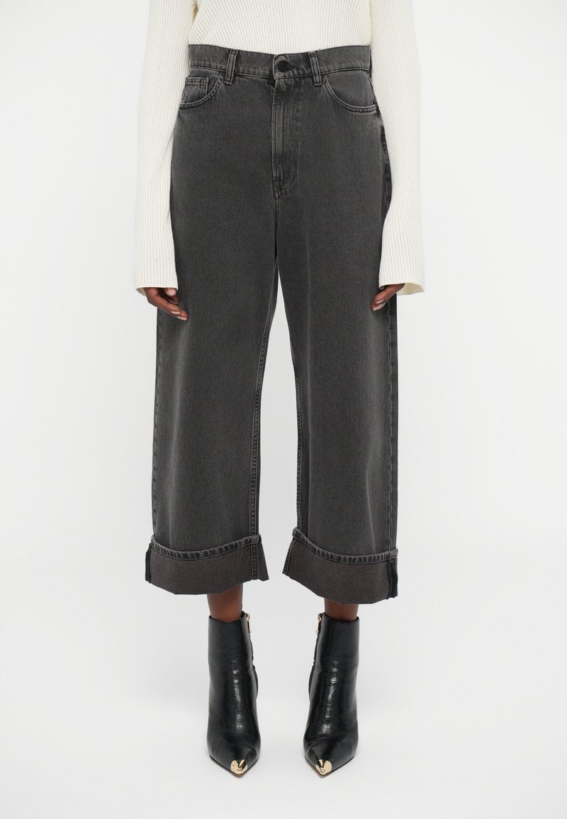 Person som har på seg høyt midjeseit, vide sorte stumpejeans med store oppklipps, en hvit langermet genser og svart spiss tå ankelstøvler.