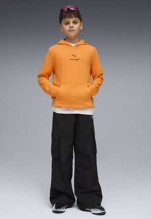 Chłopiec w pomarańczowej bluzie McLaren, czarnych spodniach cargo, czarno-białych trampkach, z różowymi okularami przeciwsłonecznymi na głowie, stoi z rękami w kieszeniach.