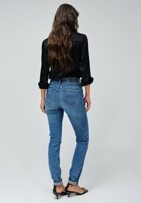 Vaqueros slim de mezclilla azul con una cintura a media altura, con bolsillos traseros y un lavado desgastado, combinados con una camiseta negra de manga larga y sandalias negras.