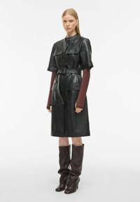 Juodas odinis suknelė su trumpomis rankovėmis, diržu juosmens srityje ir dviem priekinėmis kišenėmis. Derinama su rudo odos batais iki kelių.