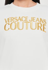 Felpa bianca con ricamo oro del testo "VERSACE JEANS COUTURE". Presenta un colletto rotondo e una trama di tessuto liscia.