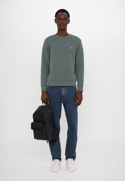 Sudadera gris con cuello redondo, jeans azul oscuro y zapatillas blancas. Lleva una mochila negra con bolsillo frontal y textura suave.