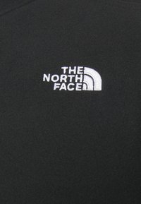 Schwarzer Fleece-Stoff mit einem weißen gestickten Logo von "THE NORTH FACE" und einem gewölbten Akzentdesign. Weiche Textur ohne sichtbare Muster.
